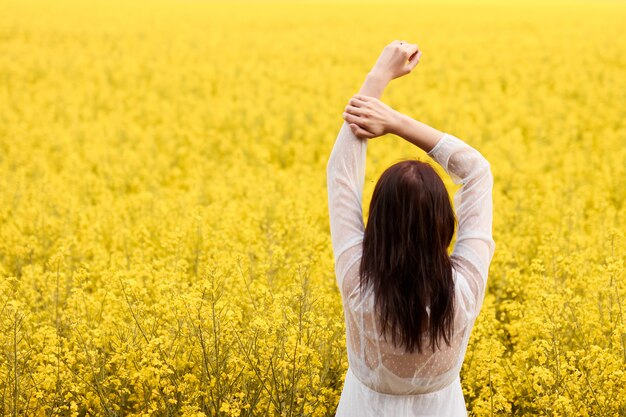 Young woman in a white dress raised her hands above a head on the yellow field with rape flowers. Concept of wedding photo in spring with copy space.