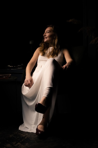 A young woman in a white dress sits relaxed on an armchair in a dark room