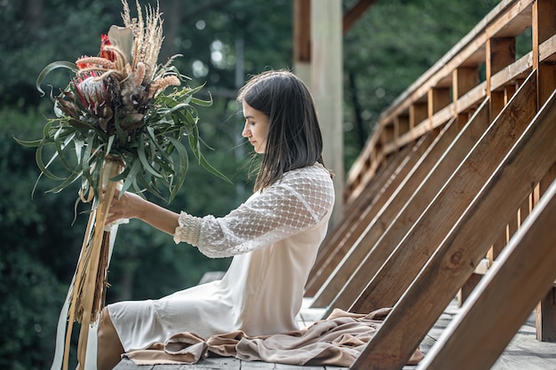 하얀 드레스를 입은 젊은 여성이 이국적인 꽃 꽃다발을 들고 나무 다리에 앉아 있습니다.