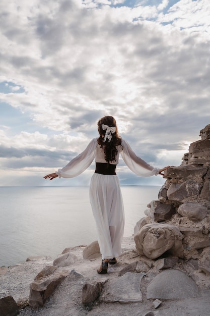 A young woman in a white dress stands barefoot on a cliff face\
in full height the dress flutters in the wind a sacred glow breaks\
through the clouds the girl looks like an angel