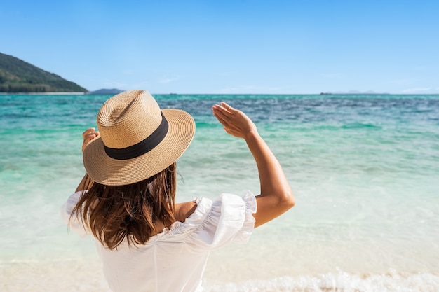 Young woman in white dress and straw hat raising her arms at the beach