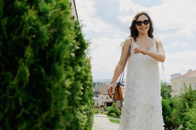 A young woman in a white dress and sunglasses poses against the background of a cottage village Good mood and summer vibe