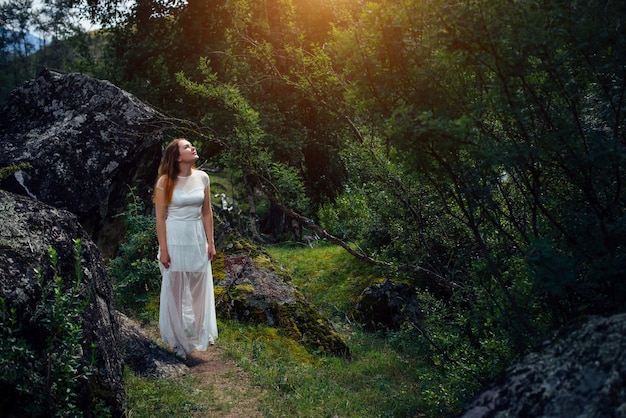 Foto giovane donna in abito bianco passeggiate nella foresta di montagna tra grandi massi grigi romantica ragazza dai capelli rossi godendo di una giornata estiva nel boschetto ombroso