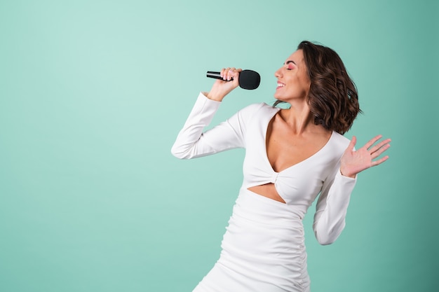 Young woman in a white evening dress on light green with bright pink makeup holds a microphone, sings karaoke