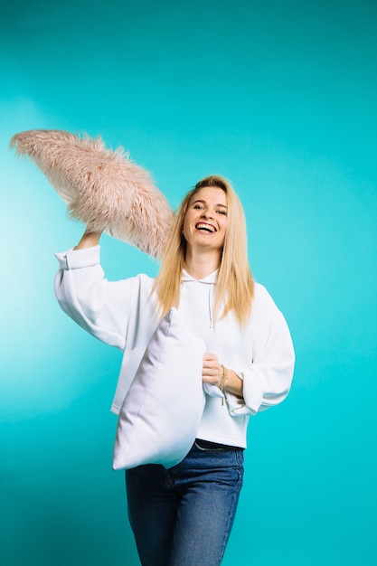 Young woman in white hoody throwing pillows on blue background. Pretty girl