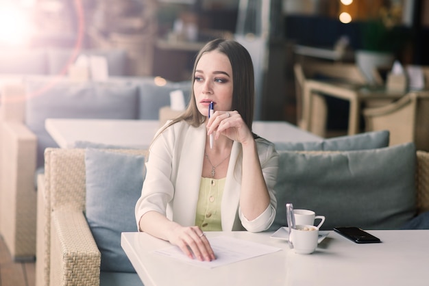 Young woman in a white jacket working in cafe with papers, drinking coffee