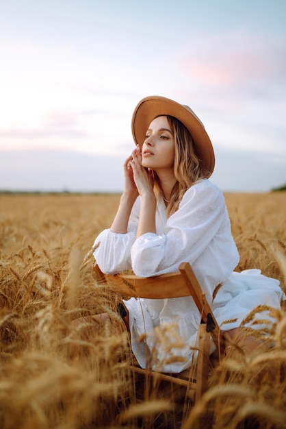 Young woman in white linen dress and hat enjoying a sunny day in a golden wheat field Summer beauty