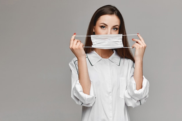 Young woman in white medical coat showing a medical protective mask and going to use it, isolate at the grey background. Copy space for your text and product. Healthcare concept