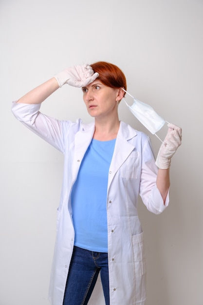 Young woman in white medical gown takes off her medical mask. Portrait of female doctor on white background. Intern finished his shift. Family doctor, tired woman therapist.