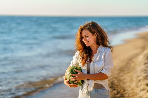 A young woman in a white shirt carries a watermelon in her hands walking along the sea on beach