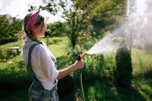 a young woman in a white shirt and denim overalls watering the lawn