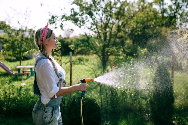 a young woman in a white shirt and denim overalls watering the lawn