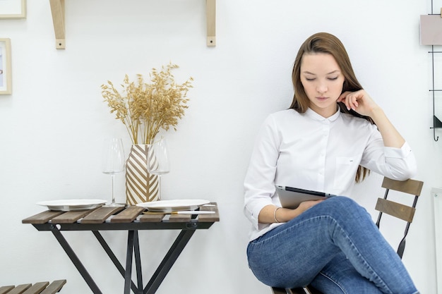 A young woman in a white shirt and jeans sits in a cafe at a table with a tablet in her hands Female office worker at lunch break Manager at a business meeting in a restaurant