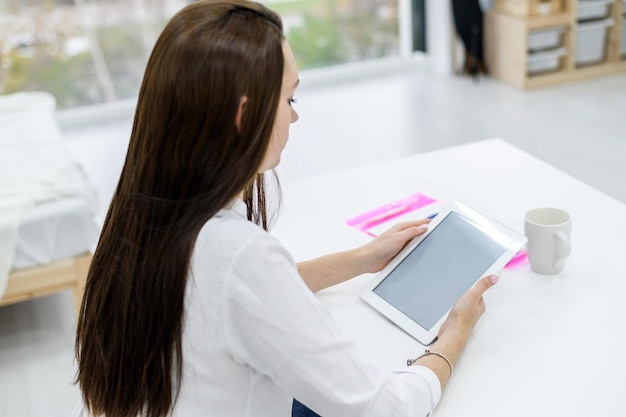 A young woman in a white shirt and jeans sits at a table with a tablet in her hands A female office manager performs work on a modern device The view from behind