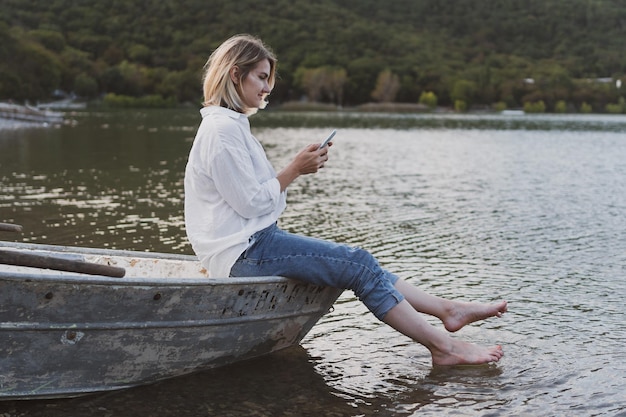 Young woman in white shirt and jeans with a phone in her hands\
sits on boat on the shore of mountain lake