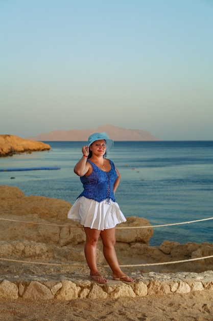 A young woman in a white skirt and a sun hat on the seashore admires nature from above.