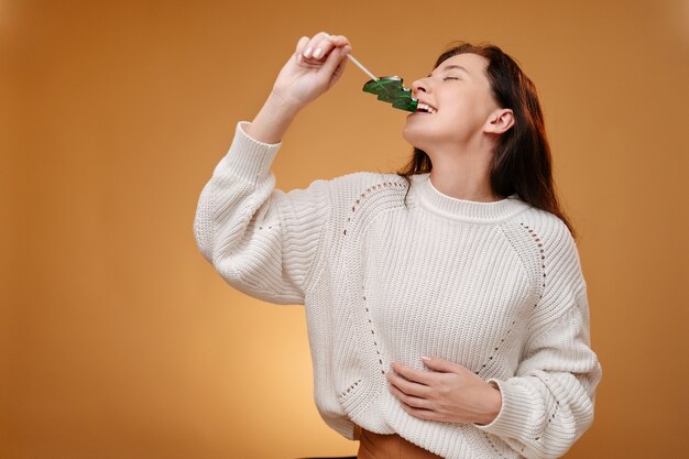 흰 스웨터를 입은 젊은 여성이 전나무 모양의 달콤한 크리스마스 롤리팝을 맛본다