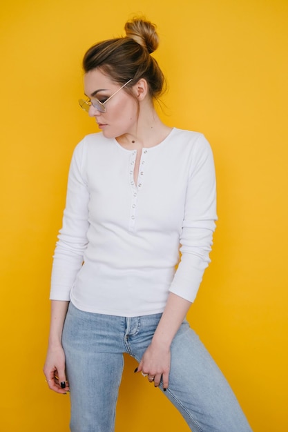 Photo a young woman in a white sweatshirt and blue jeans stands in the studio against a yellow background