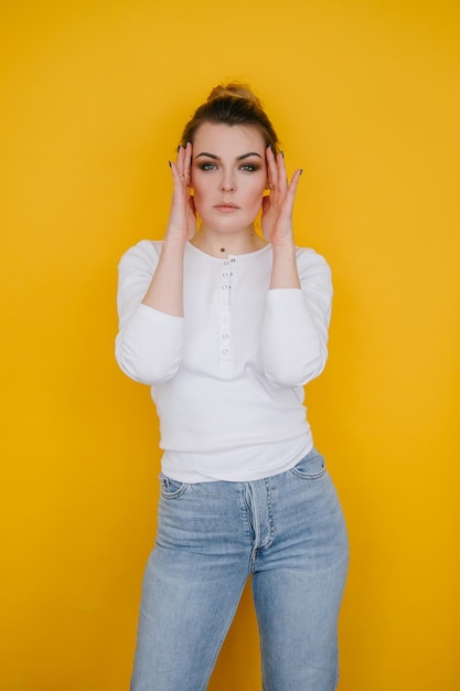 A young woman in a white sweatshirt and blue jeans stands in the studio against a yellow background