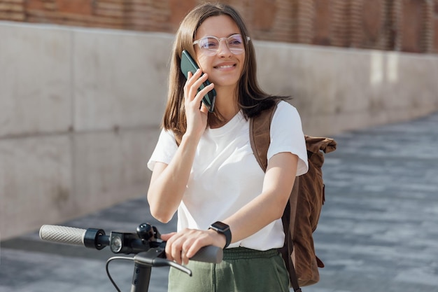 Young woman in white t shirt standing on electric scooter and talking to smartphone on urban street