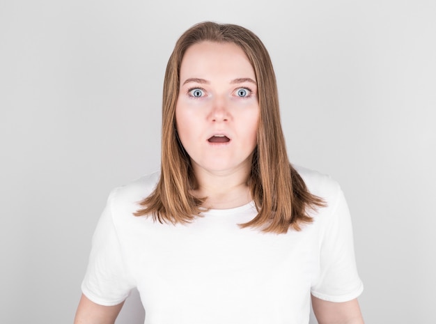 Young woman in a white T-shirt stands against a gray wall in great surprise.