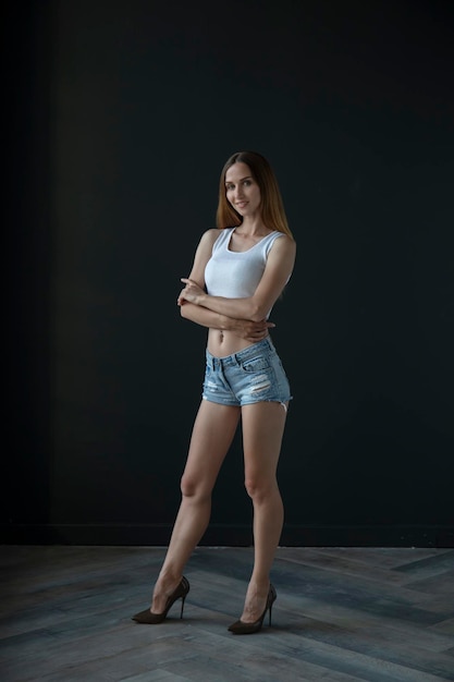 young woman in a white tank top and denim shorts on a dark background, full length portrait