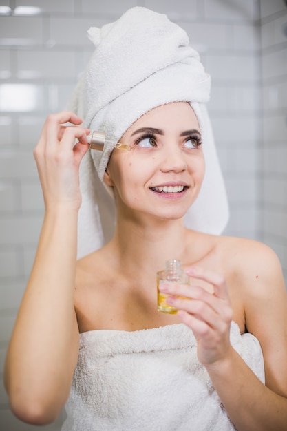 Young woman in white towel apply moisturising oil on face skin. Self care, beauty and skin care concept.