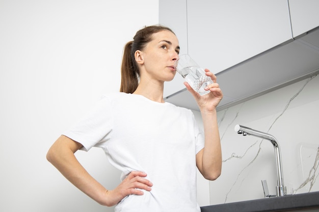 Young woman in a white tshirt in the kitchen drinks water from a glass cup