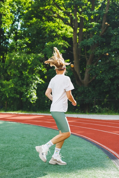 Молодая женщина в белой футболке и кроссовках с кепкой на голове Она разминается перед тренировкой на открытом воздухе на стадионе Здоровый образ жизни