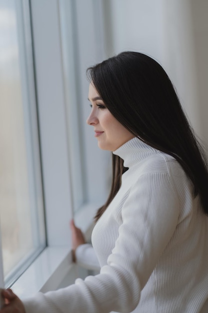 Молодая женщина в белой водолазке смотрит в окно, думая или мечтая