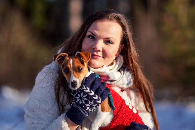 Young woman in winter jacket holding her Jack Russell terrier on hands blurred trees background