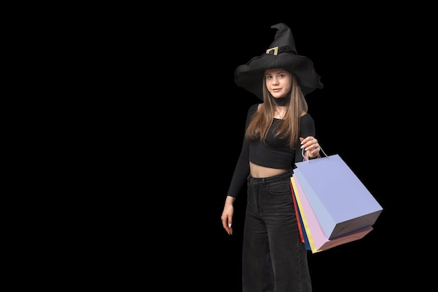 Young woman in witchs hat with colorful paper shopping bags in hands Copy space Black Friday concept Isolated on black background