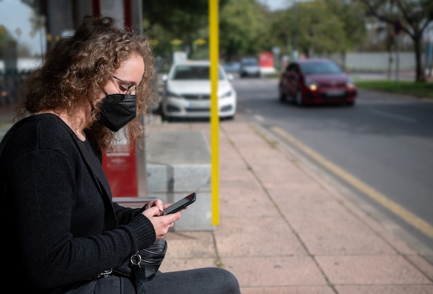 Фото Молодая женщина в маске сидит на автобусной остановке и печатает на своем смартфоне