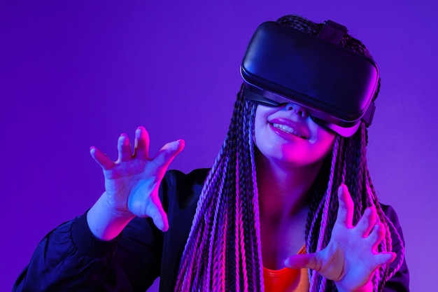 Young woman with afro braids using vr glasses on dark purple neon background