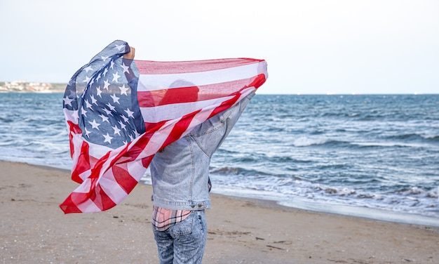 A young woman with an American flag runs by the sea. The concept of patriotism and independence day celebrations.