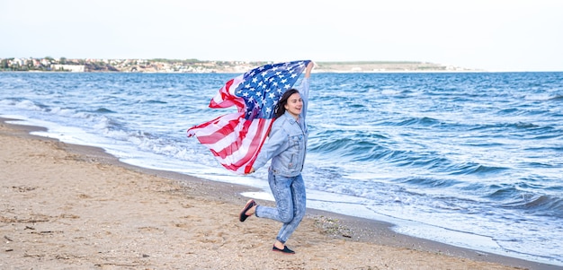 アメリカの国旗を持つ若い女性が海を走っています。愛国心と独立記念日のお祝いの概念。