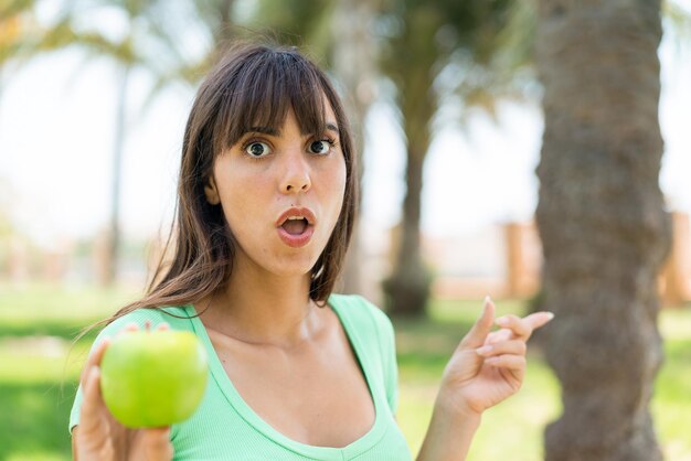 사진 야외에서 사과를 들고 있는 젊은 여성이 놀라고 측면을 가리키고 있습니다.