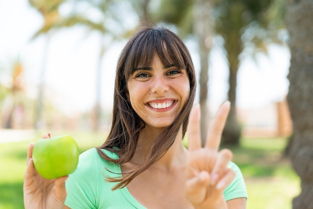 Foto giovane donna con una mela all'aperto sorridente e mostrando segno di vittoria