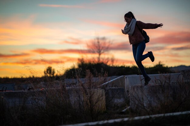 Foto giovane donna con le braccia tese in piedi su un muro di pietra contro il cielo durante il tramonto.