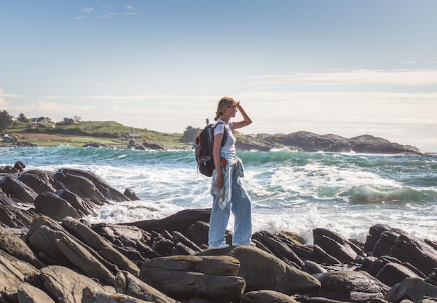Молодая женщина с рюкзаком стоит на камне и смотрит на океанские волны