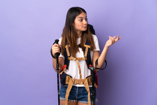 Young woman with backpack and trekking poles isolated on purple background extending hands to the side for inviting to come