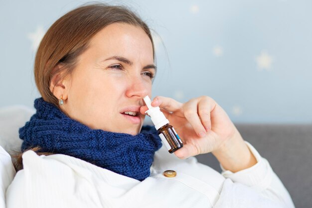 Photo young woman with bad cold lying in bed under blanket and using nasal drops