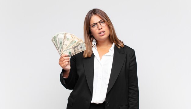 Young woman with banknotes feeling puzzled and confused, with a dumb, stunned expression looking at something unexpected