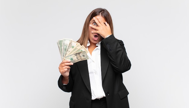 Young woman with banknotes looking shocked, scared or terrified, covering face with hand and peeking between fingers