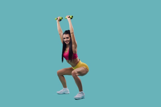 Young woman with beautiful body performs dumbbell squats isolated on blue background with copy space.