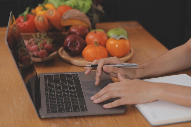 A young woman with a beautiful face in a blue shirt with long hair eating fruit sitting inside the kitchen at home with a laptop and notebook for relaxation Concept Vacation