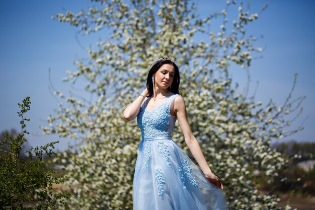 Young woman with beautiful make-up with a vintage hoop in a blue light dress with a bouquet posing on a background of green foliage in a park. Fresh portrait attractive girl with white flowers.