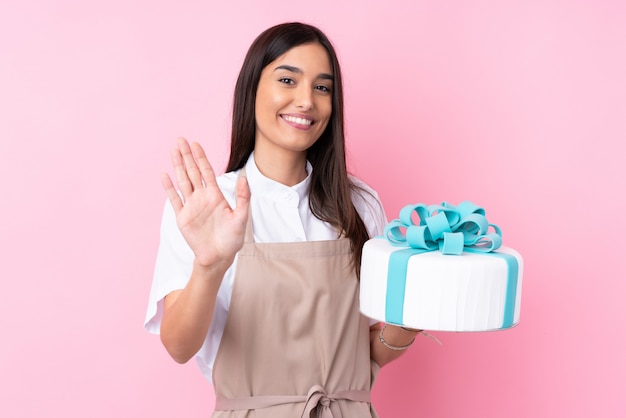 Giovane donna con una grande torta sul muro isolato salutando con la mano con espressione felice