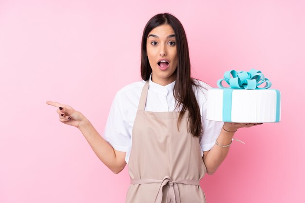 Young woman with a big cake over surprised and pointing finger to the side