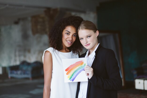 Young woman with blond hair in black suit holding lgbt sign in hand with pretty african american woman with dark curly hair in white dress while dreamily  together on wedding ceremony